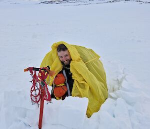 Image of Expeditioner crouching inside of yellow bivvy bag. Foreground displays broken-up snow. Background has a dark grey cloudy sky with sun glow.