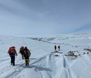 Image of two pairs of expeditioners with survival packs on, walking away from the camera conducting training. The foreground is a snow dotted with rocks. Background is a flat blue sky with snow cliffs.