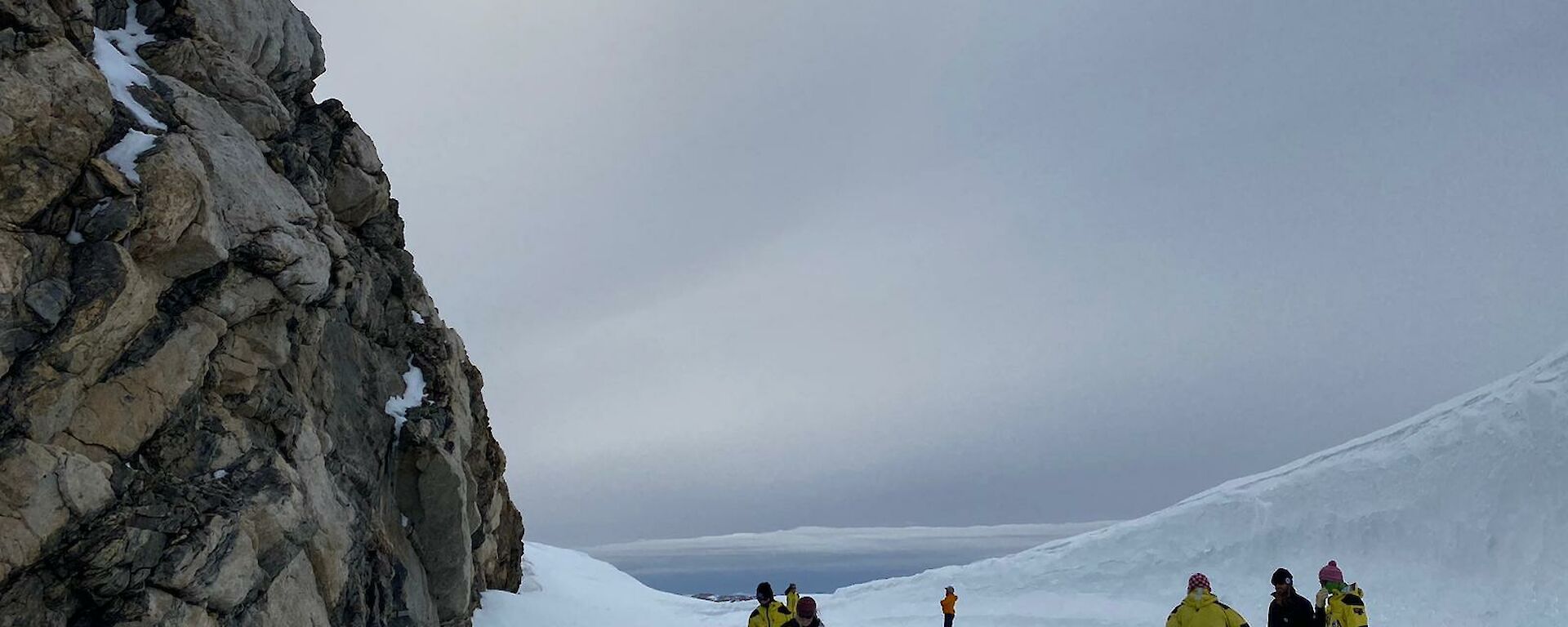 Image of Tiger Rock to the left of the image. Sun with halo glow is in the middle to top of the image. The front of the image shows flat ice that surrounds the rock with Expeditioners walking on the ice. To the right is a wave-shaped structure of snow.