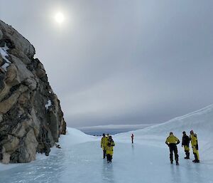 Image of Tiger Rock to the left of the image. Sun with halo glow is in the middle to top of the image. The front of the image shows flat ice that surrounds the rock with Expeditioners walking on the ice. To the right is a wave-shaped structure of snow.