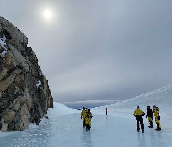 Image of Tiger Rock to the left of the image. Sun with halo glow is in the middle to top of the image. The front of the image shows flat ice that surrounds the rock with Expeditioners walking on the ice. To the right is a wave-shaped structure of snow.
