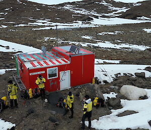 A red hut sits on a rocky hill surrounded by snow with six people in yellow in the foreground