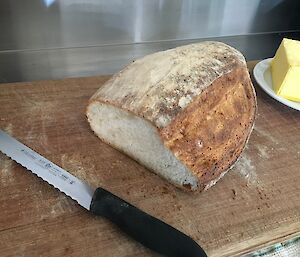 A cut loaf of sourdough on a wooden bread board with bread knife and butter