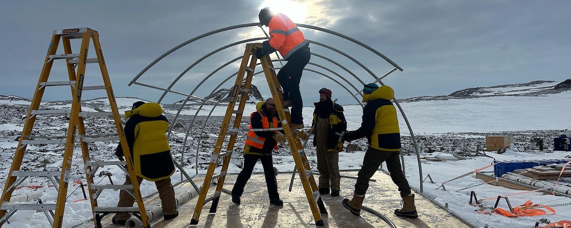 Five people in hi vis building the fame of a tent on a platform in a snowy landscape