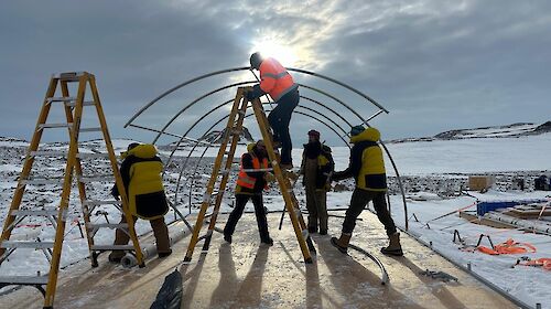 Five people in hi vis building the fame of a tent on a platform in a snowy landscape