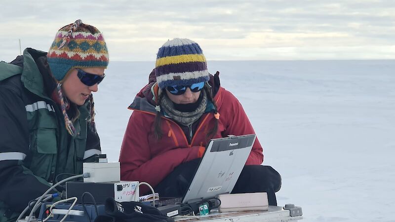 Two women in winter woollies look at a computer on an icy plain