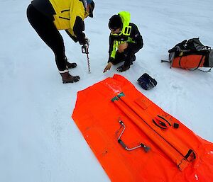 A red box sits on the ice while two people in yellow put a jagged thin instrument through the ice