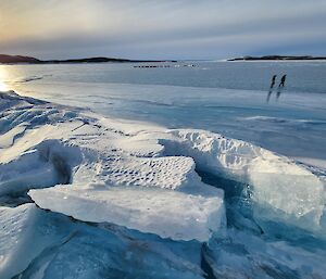 Two figures in the distance on an icy lake with ice chunks in the foreground