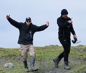 Two men walking, one has arms up and looks to camera other is talking on radio