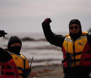 Close up of two boat crew, wearing life jackets, dry suits and beanies, face camera and hold arms up as if showing muscles