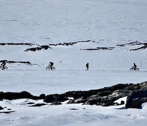 Image of three people on fat-tyre bikes and one person running on the Ski Loop. The people are framed by rocks and snow in the fore and background.