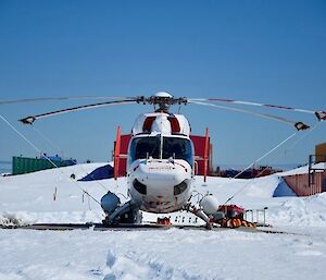 Image of white BK117 Helicopter centres the image. White snow in the foreground, bright blue sky as the backdrop.
