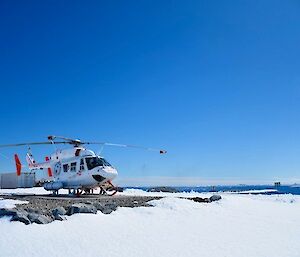 Image of a white BK117 Helicopter to the left side of the image. Blue sky and water fill the background with beaming white snow contrasting in the foreground.