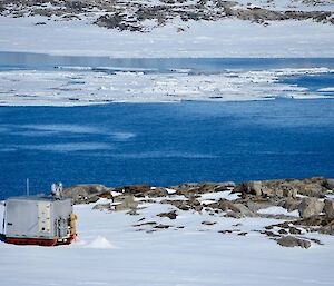 Image of Silver Chalet: a metal shipping container converted to a hut. Foreground has snow and grey rocks. The mid-ground has a deep blue body of water with icer bergs clustered together. The background is distant snow with rocks.