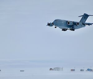 Image of a RAAF C17 aircraft coming in for landing. The background of the image features small colourful buildings with drifting snow creating haze.