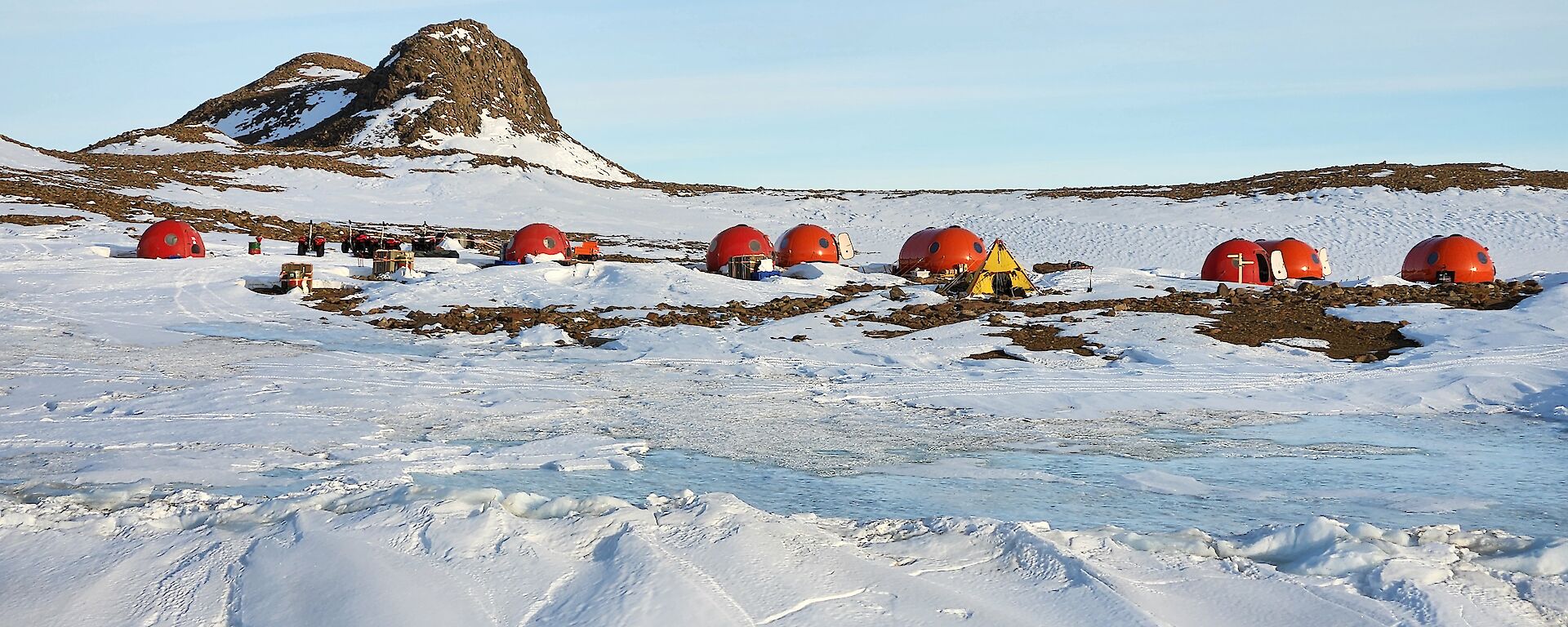 A group of seven semi-circular red fibreglass huts set up on snow, with a rocky peak behind.