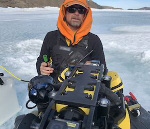 A man in an orange hat standing on a frozen over lake, with a small, submersible vehicle that can be lowered through a hole in the ice.