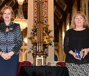 A woman with red hair and blue suit on the left, Alison dean in a black top and skirt, holding the Antarctic Medal.