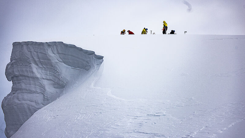 A snow cliff to the left of frame with a group of people to the right at the top, doing training