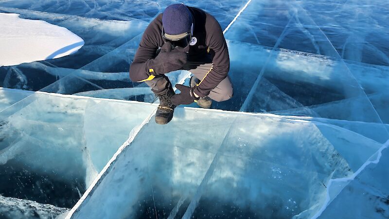 A person crouches down, staring into a frozen lake with lots of fault lines in it
