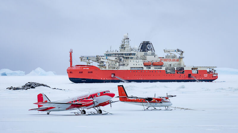 Two planes stand on the ice in front of a big red icebreaker