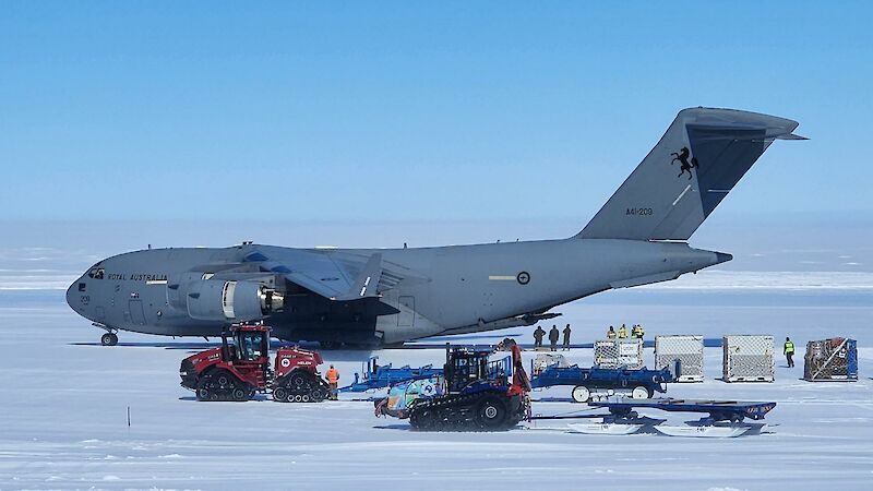 A massive grey plane stands on an ice runway with lots of heavy plant equipment around it, unloading