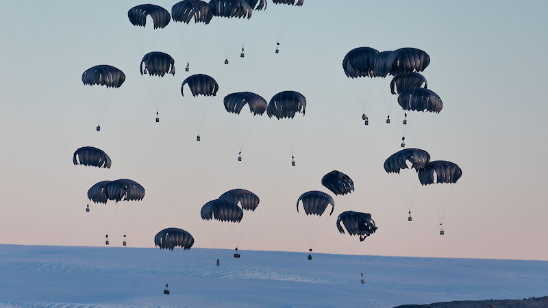 More than a dozen black parachutes with loads attached float down to the snow.