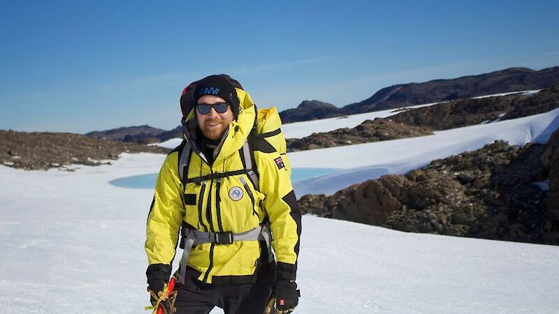 A man in yellow stands on a snowy slope with hills in the background