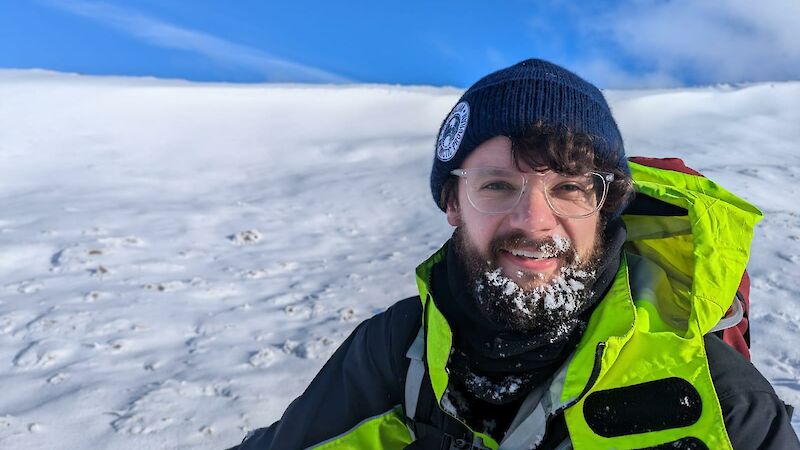A man in yellow with snow in his beard, stands on a snowy slope under blue sky
