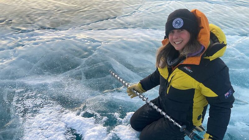 A woman in yellow Antarctic gear drills a hole in the ice