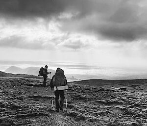 Black and white photo of two people walking along a ridge with rugged hills, sea and clouds in front of them.