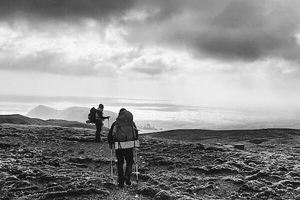 Black and white photo of two people walking along a ridge with rugged hills, sea and clouds in front of them.