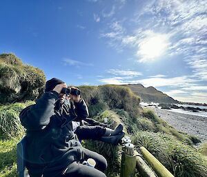 A person with binoculars looks out from a grassy hill out to the sea