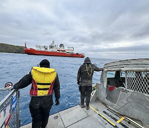 Two people stand on a metal flat-platform boat looking over to a big red icebreaker