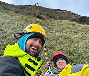Two people smiling holding a rope that leads up a grassy hill, with people at the top
