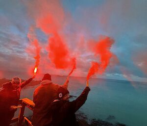 A group of people letting off flares at dusk with the sea spread out in front of them