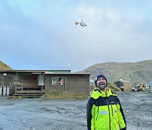 A smiling man in yellow on a grey gravel flat are with helicopter above