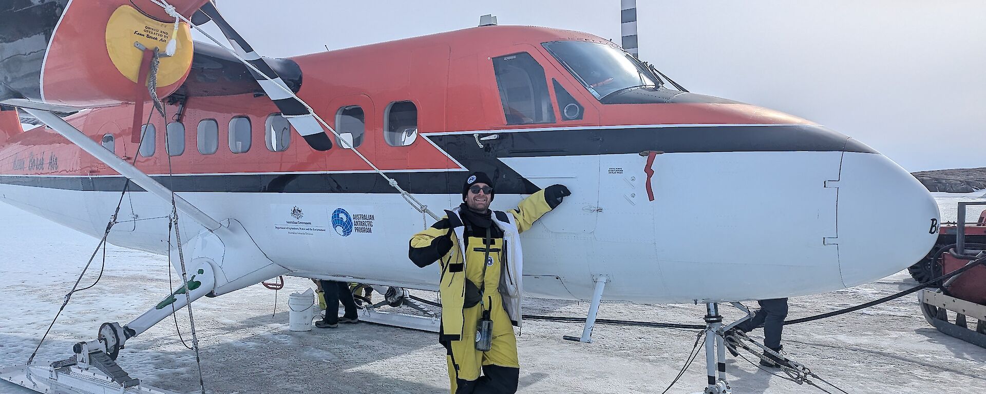 An expeditioner in yellow outers standing next to a Twin Otter place on the sea-ice in front of Mawson Research station