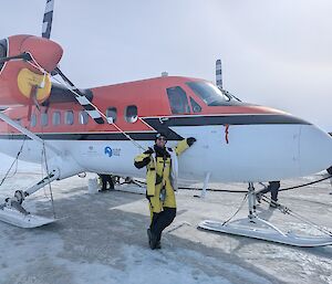 An expeditioner in yellow outers standing next to a Twin Otter place on the sea-ice in front of Mawson Research station