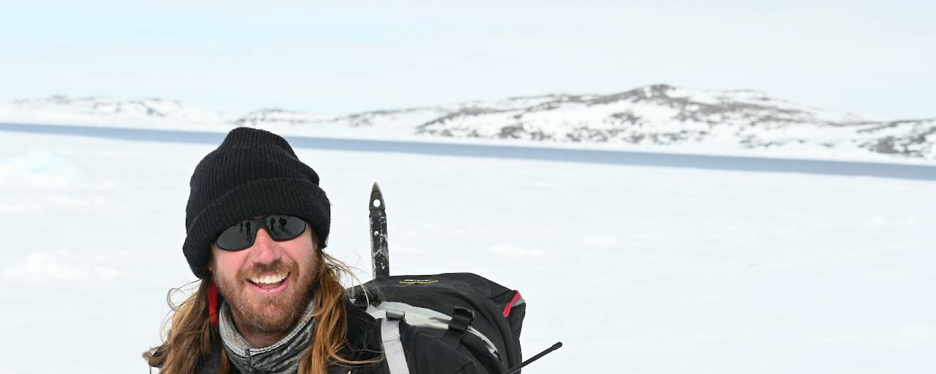 Image of expeditioner smiling at camera. Background contains snowy mountains with a pale blue sky.