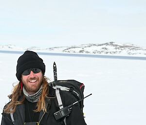 Image of expeditioner smiling at camera. Background contains snowy mountains with a pale blue sky.