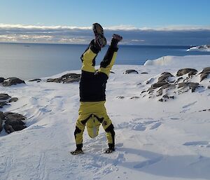 Image of Expeditioner in yellow survival suit doing a handstand in the snow. Foreground is snow. Background is a blue sky and deep blue water meeting the sky at the horizon line.