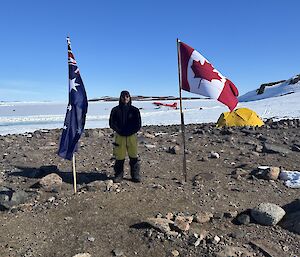 Image of Expeditioner standing between Australian and Canadian flags. Foregound is rock. Background is snow and sky with a red and white Basler aircraft.