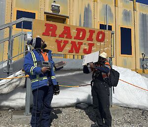 Image of ITO and CTO holding communications equipment. Personnel are standing infront of the yellow Operations Building with 'Radio VNJ' sign behind them.