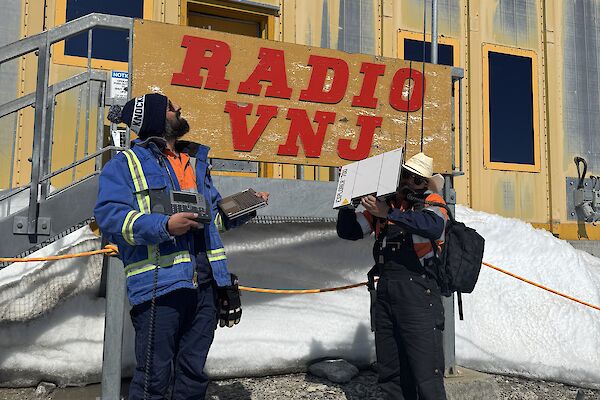 Image of ITO and CTO holding communications equipment. Personnel are standing infront of the yellow Operations Building with 'Radio VNJ' sign behind them.