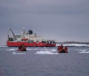 A large red ship with 2 small boats in front with some floating small bergy bits