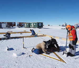 Three people creating a triangular structure in the snow, inside a wooden frame.