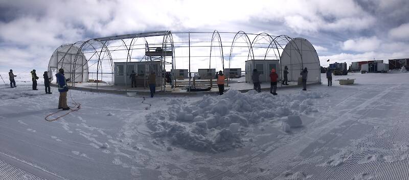 A cylindrical metal frame, about 27 metres long, rises above a compacted snow floor, in Antarctica. People stand on one side preparing to haul a tarpaulin over the structure using ropes.