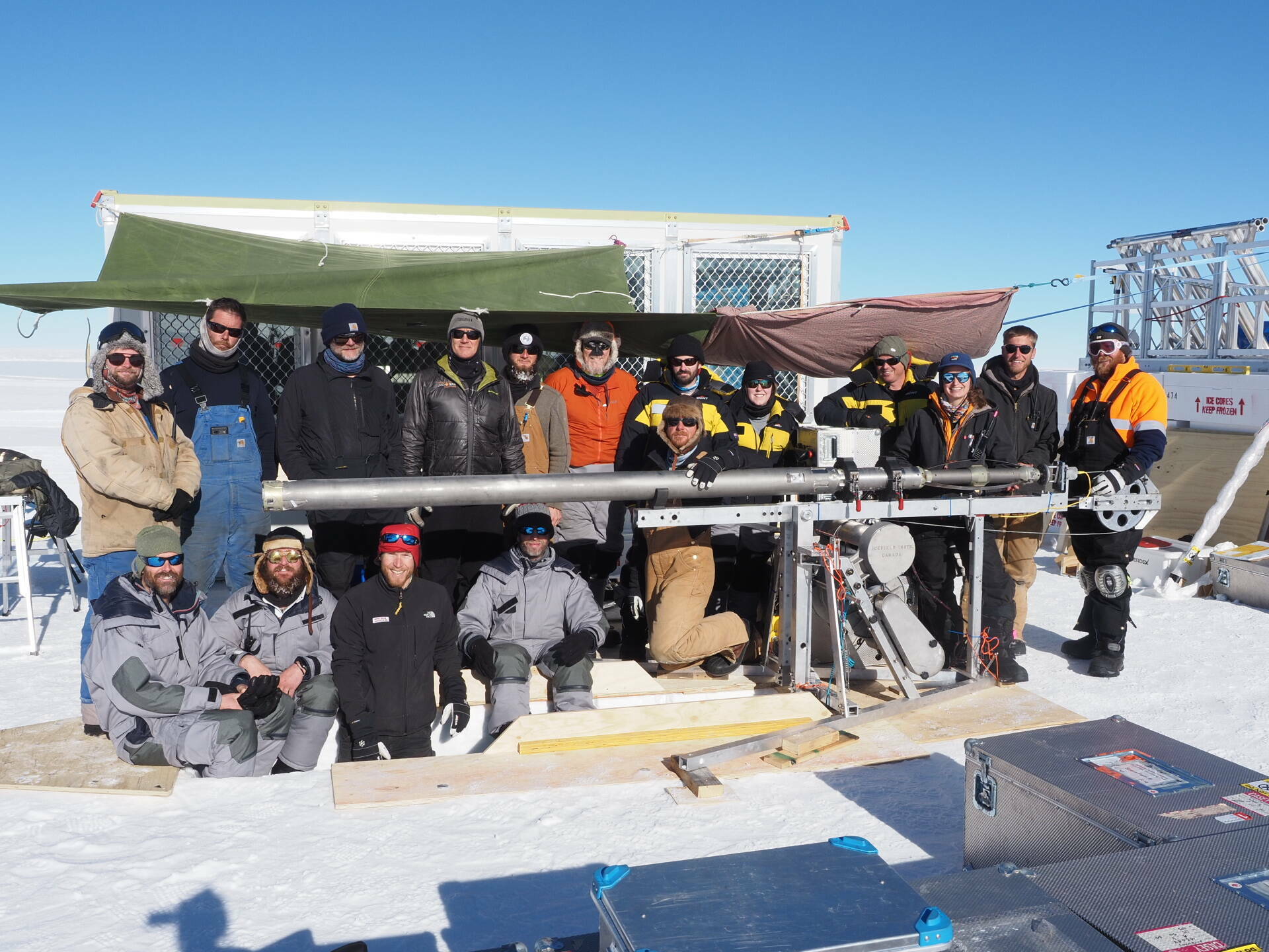 A team of 16 men and women standing around a long metal drill and drilling trench.