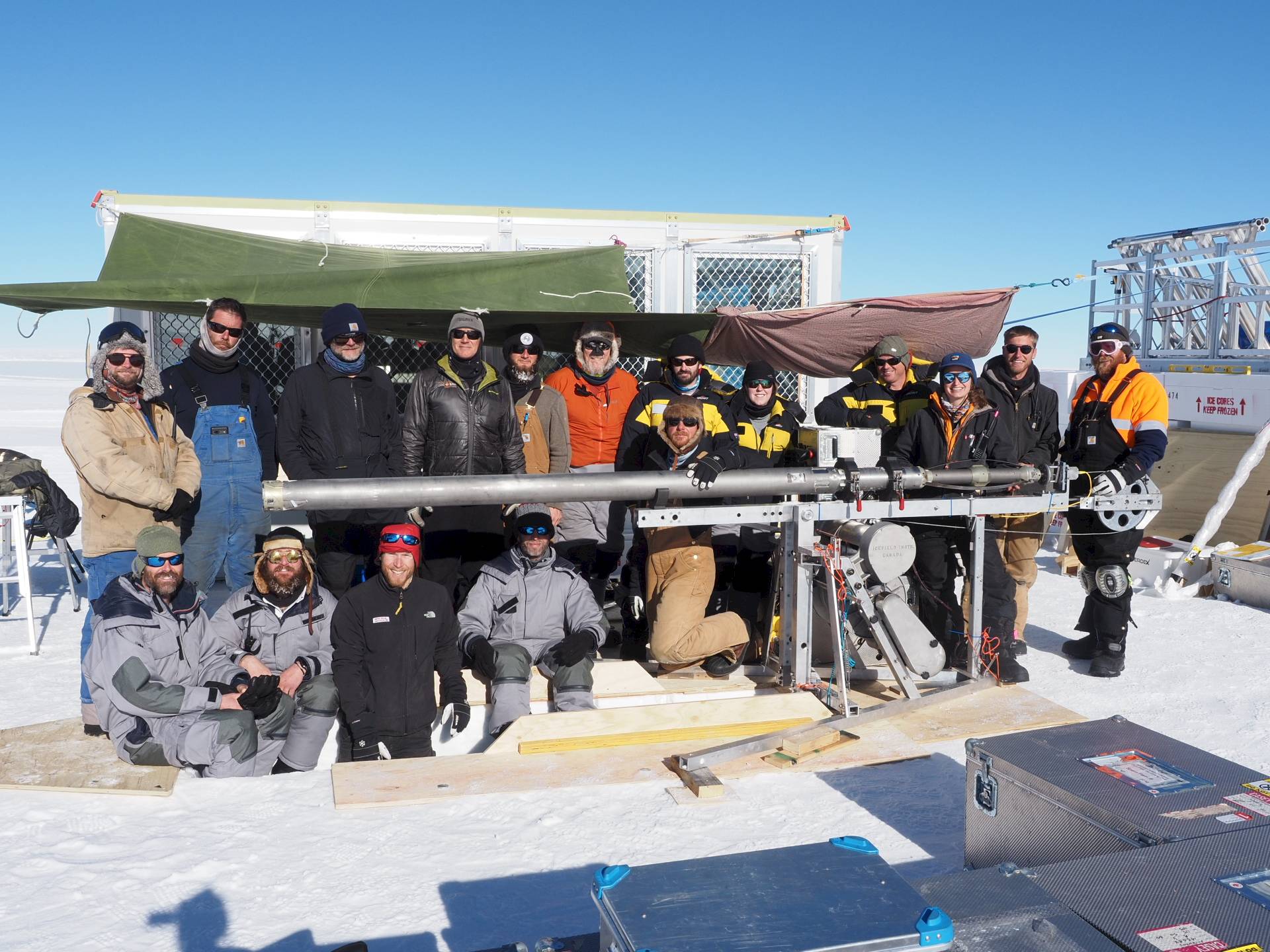 A team of 16 men and women standing around a long metal drill and drilling trench.